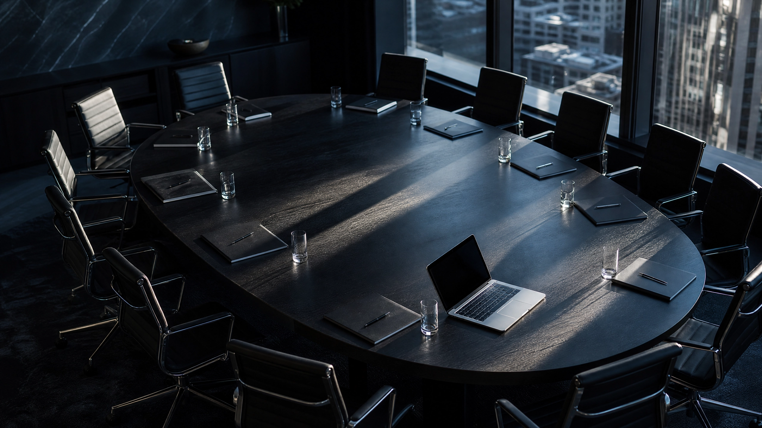 Empty oval boardroom table with notepads, water glasses, and a single open laptop — the aftermath or anticipation of a high-stakes conversation