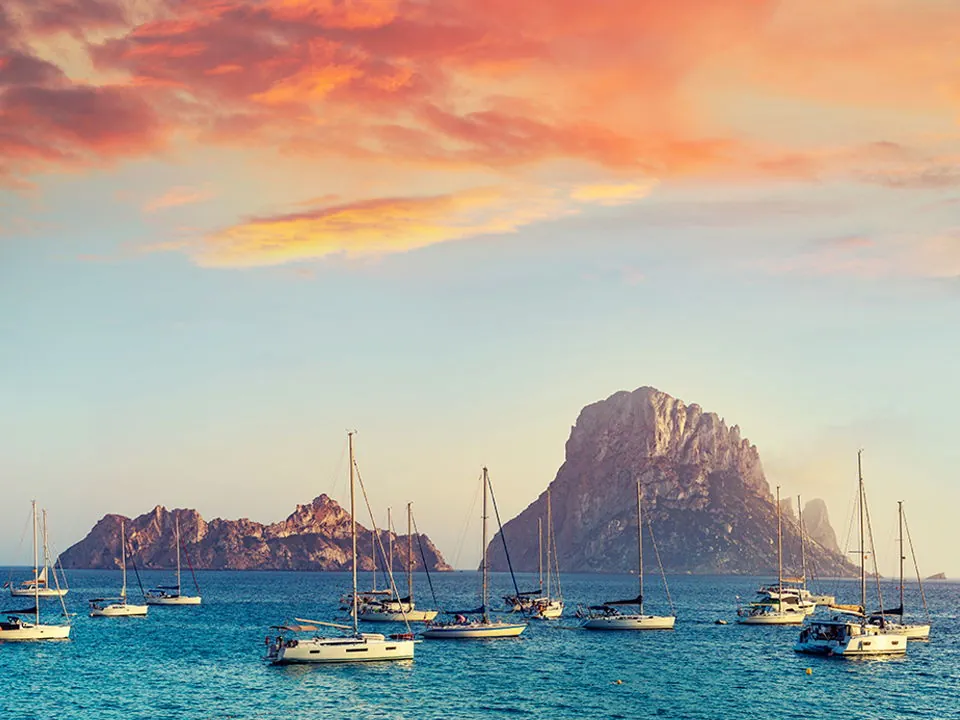 Sailing yachts anchored at sunset near Es Vedrà, Ibiza, in a marina setting.