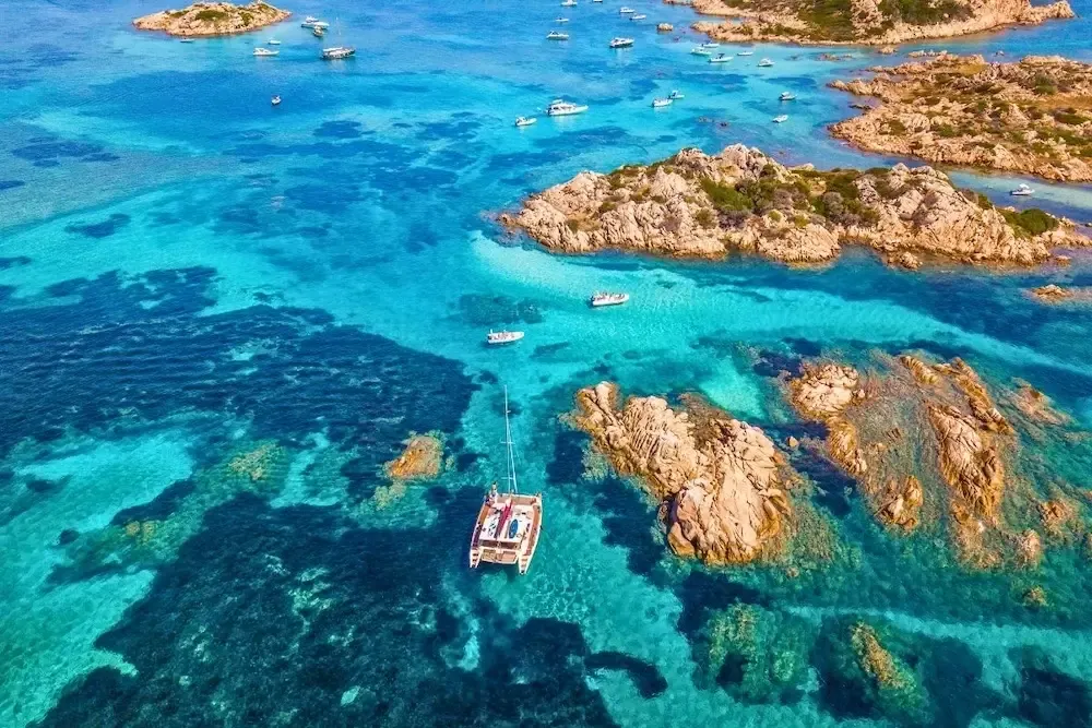 Aerial view of a catamaran and boats in turquoise water in the La Maddalena Archipelago, Sardinia.
