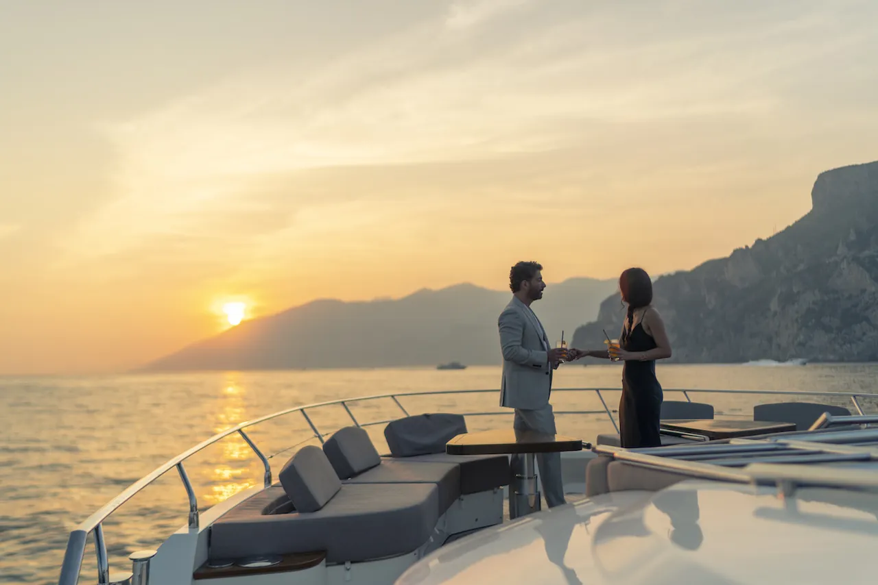 Guests in evening dress on a yacht deck at sunset with a mountainous coastline behind