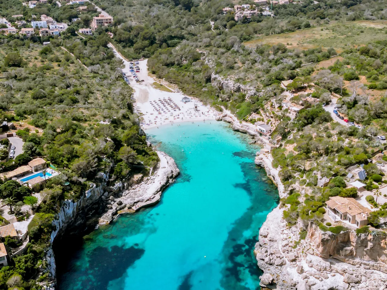 Aerial view of the turquoise cove and sandy beach at Cala Pi, Mallorca, with people in the distance.