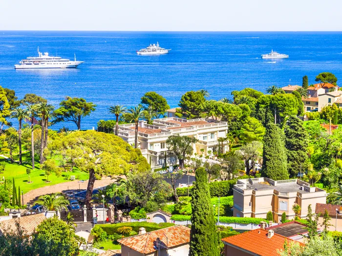 Aerial view of a white villa and formal gardens overlooking the French Riviera, with superyachts anchored offshore.