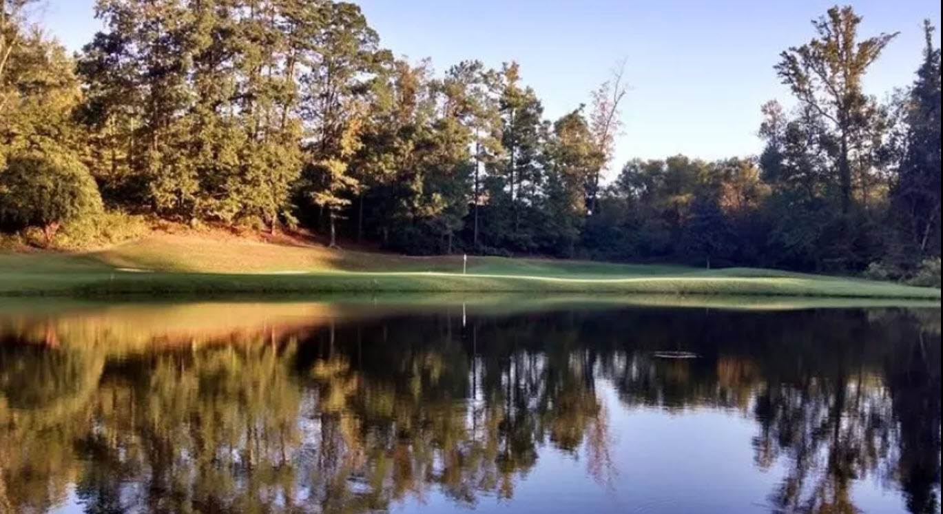 Panoramic view of a lush green golf course fairway with mature trees and a distant clubhouse under a clear sky at Duke University Golf Club