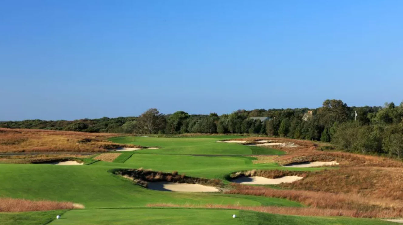 Panoramic view of the iconic Shinnecock Hills Golf Club clubhouse and a links-style hole with fescue grasses under a clear sky.
