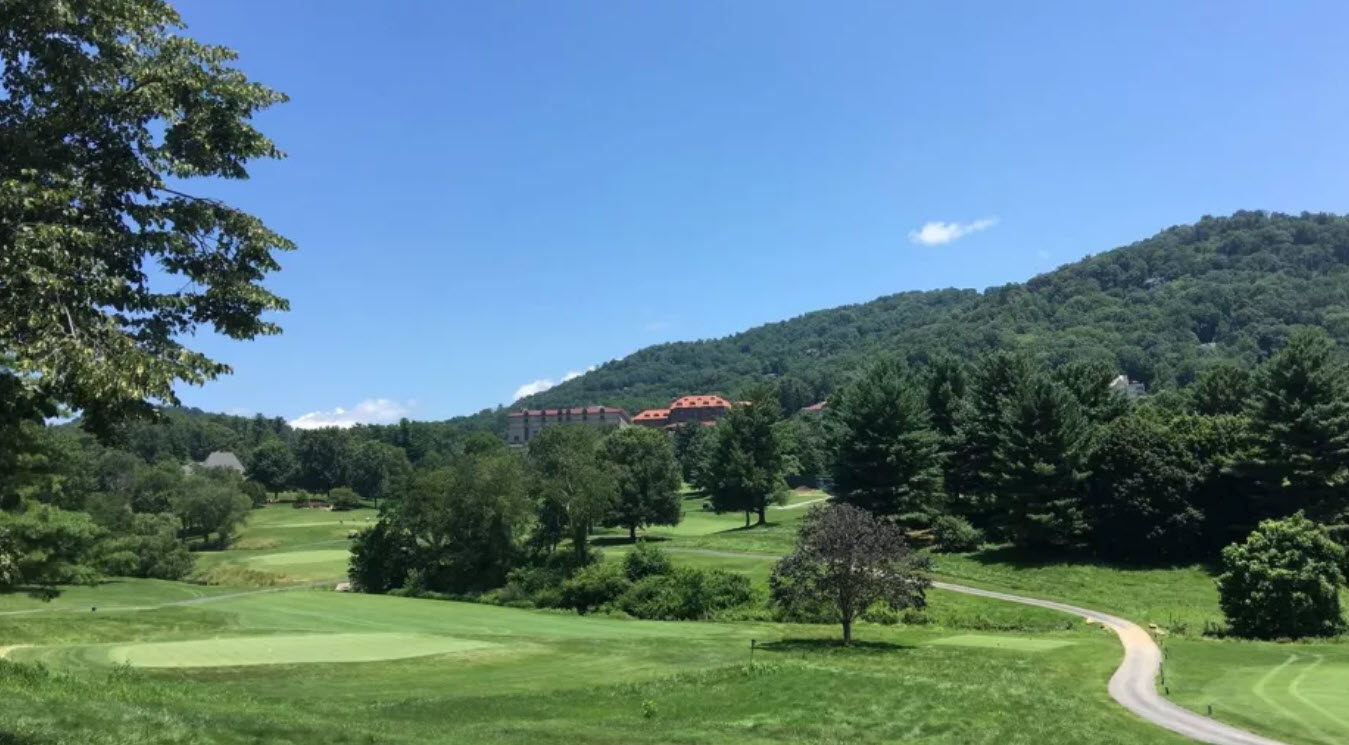 Aerial view of The Omni Grove Park Inn Golf Course with golfers on a green, surrounded by lush trees and distant Blue Ridge Mountains.