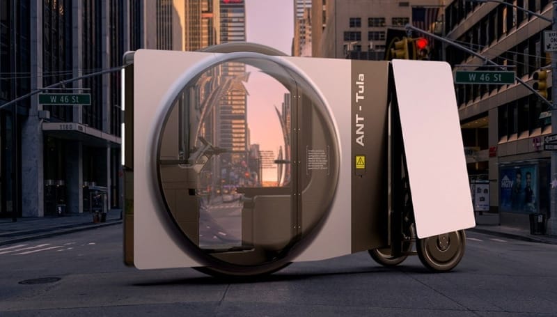 Futuristic compact vehicle with a rounded glass cabin and large rear door, parked in an urban street setting with tall buildings and traffic lights in the background.