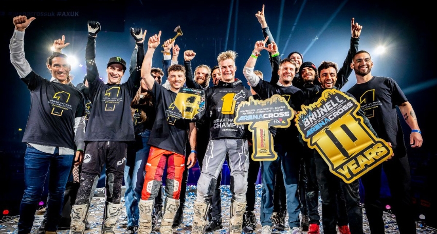A group of motocross racers and supporters celebrate on stage, holding victory signs and trophies, with joyful expressions and raised arms.