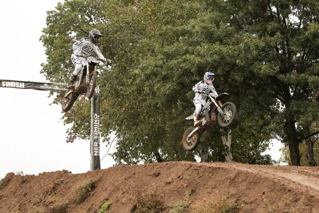 Two dirt bikers performing an impressive jump on a dirt track.
