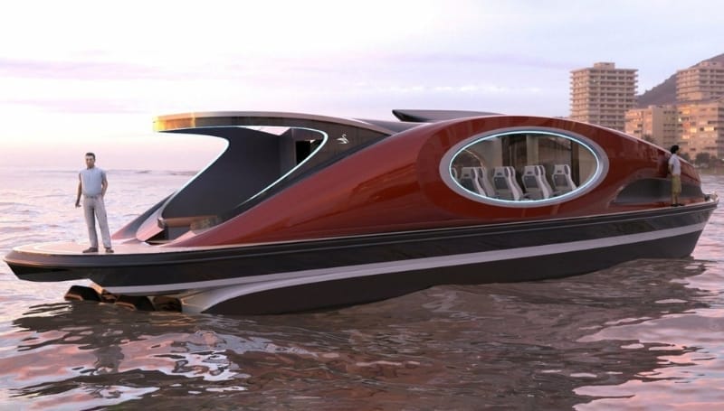 A modern red-and-black boat with large oval windows is anchored near a shoreline at sunset. A man stands at the boat's stern. Apartment buildings are visible in the background.