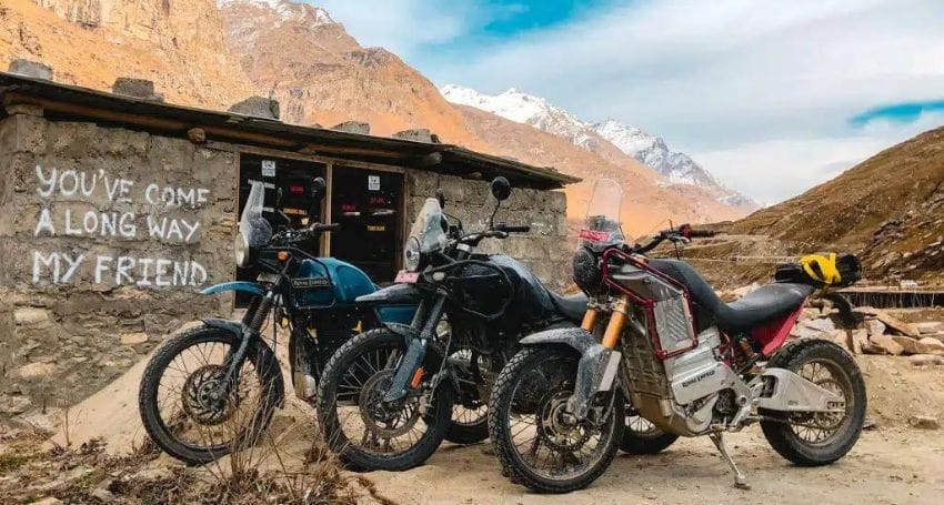 Three Royal Enfield Himalayan motorcycles parked in front of a rustic building with the message "you've come a long way my friend" painted on its wall, set against a backdrop of mountains and a