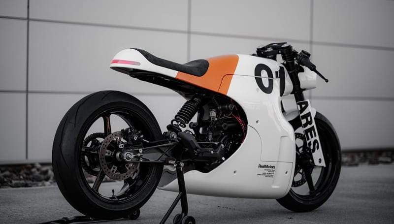 A white and orange racing motorcycle is parked on a stand, set against a minimalist grey wall background.