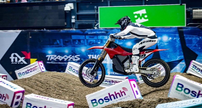 A person in motocross gear rides a red dirt bike on an indoor track with multiple sponsor banners in the background.
