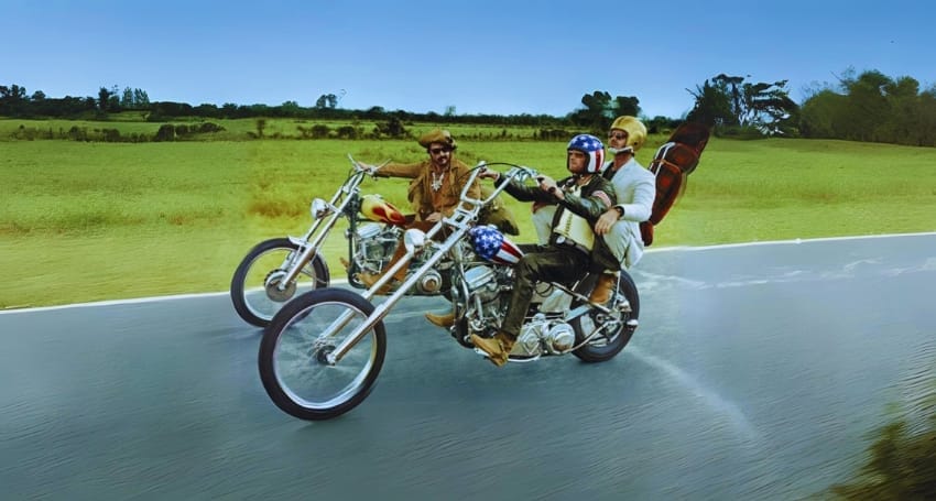 Two motorcyclists riding chopper bikes on a road, one carrying a guitar on his back.