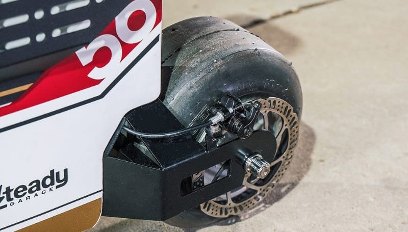 Close-up of the rear wheel and brake mechanism of a racing scooter on a concrete surface. The wheel, appearing worn with visible treads, showcases the gritty endurance of Moto Distracto. Part of the scooter frame and a partial number "59" are also visible.