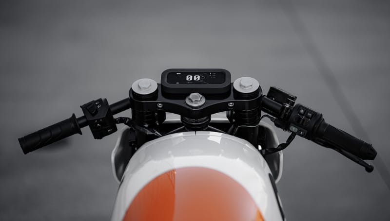 Close-up of motorcycle handlebars featuring modern digital display and control buttons. The motorcycle has a sleek, white and orange design.