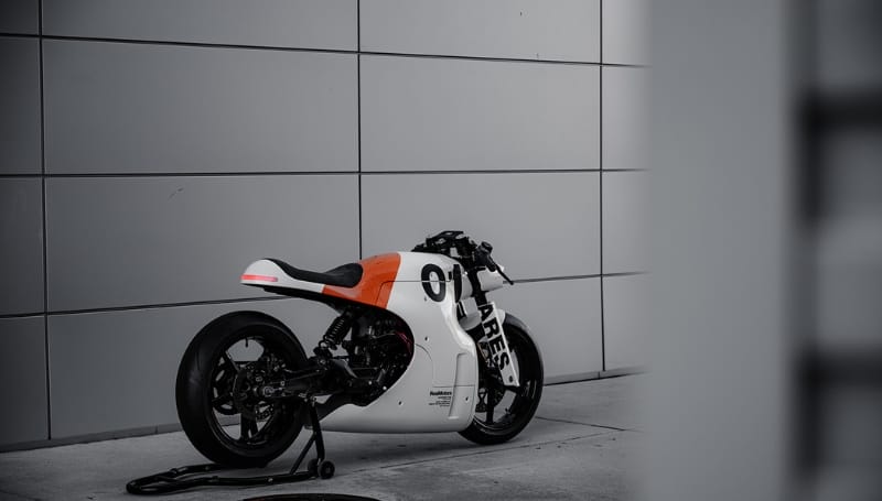 A custom white and orange racing motorcycle is parked on a concrete surface in front of a modern, steel-paneled wall. The motorcycle is on a stand, with the number "01" visible on its body.