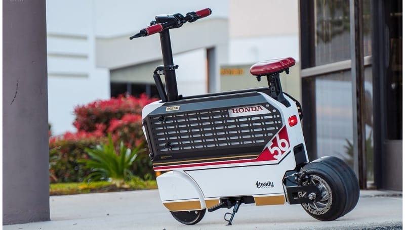 A compact Honda Motocompacto electric scooter with a white and black design, red accents, and the number 36 on the side, set against an urban background with plants and buildings.