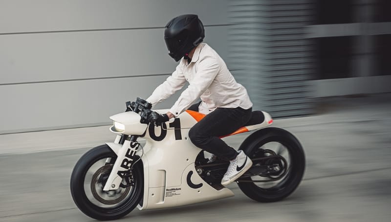 A person in a white shirt, black helmet, and jeans rides a white motorcycle with black and orange accents on a concrete surface.