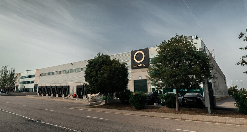 A large white industrial building labeled "Stark" with trees and a few parked cars in front, beside a wide empty street under an overcast sky.