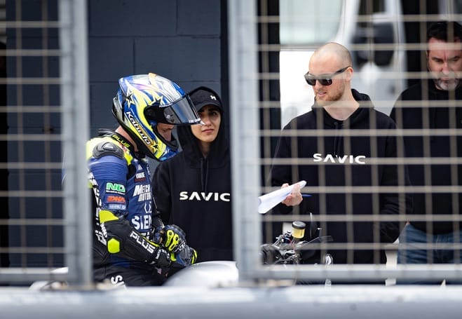 A group of people standing next to a Savic Motorcycle during high-speed tests.