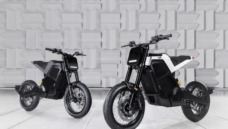 Two modern luxury electric motorcycles, one black and one white, parked side by side in front of a geometrically patterned gray wall.