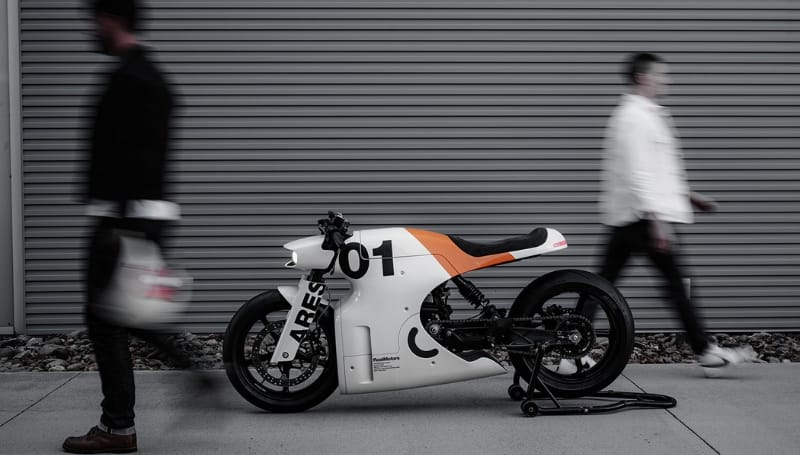 A stationary custom motorcycle labeled "01" and "ARES" is parked on a stand against a metal-paneled wall, with two blurry pedestrians walking past it.