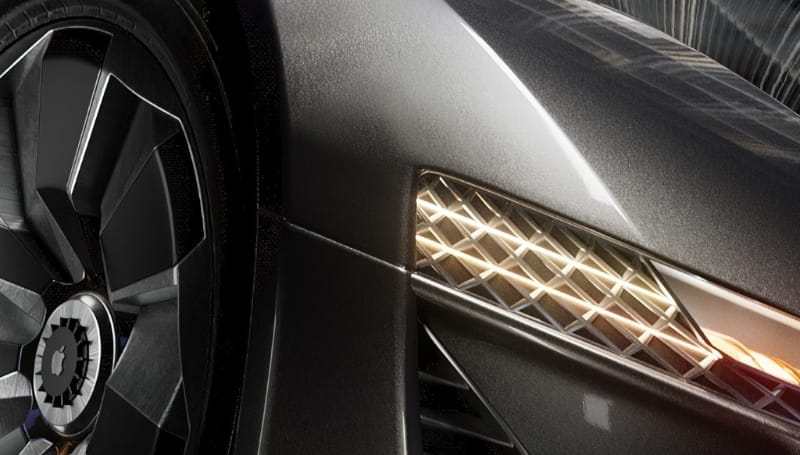 Close-up of a modern car's front quarter panel, showcasing a detailed wheel design and intricate headlight pattern. The car's metallic gray paint reflects the surroundings.