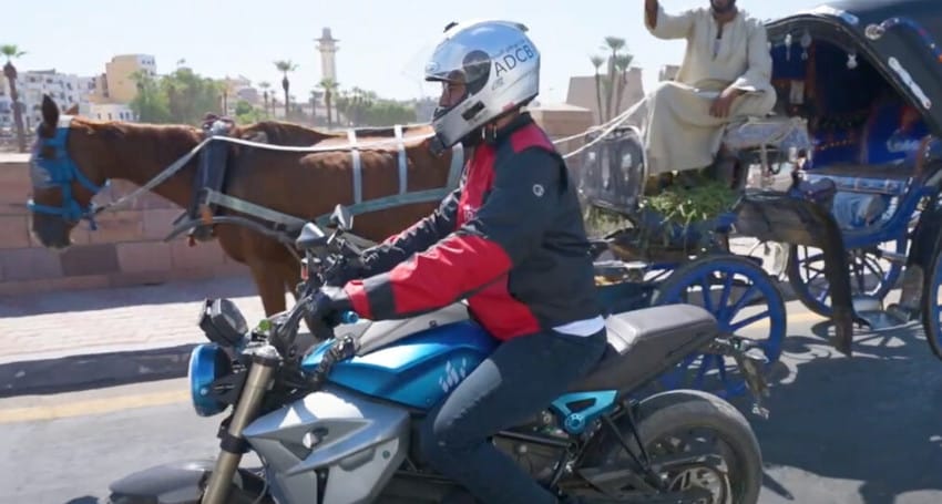 A motorcyclist, wearing a helmet and red jacket, rides past a horse-drawn carriage on a city street, showcasing the growing presence of electric motorcycles in Egypt.