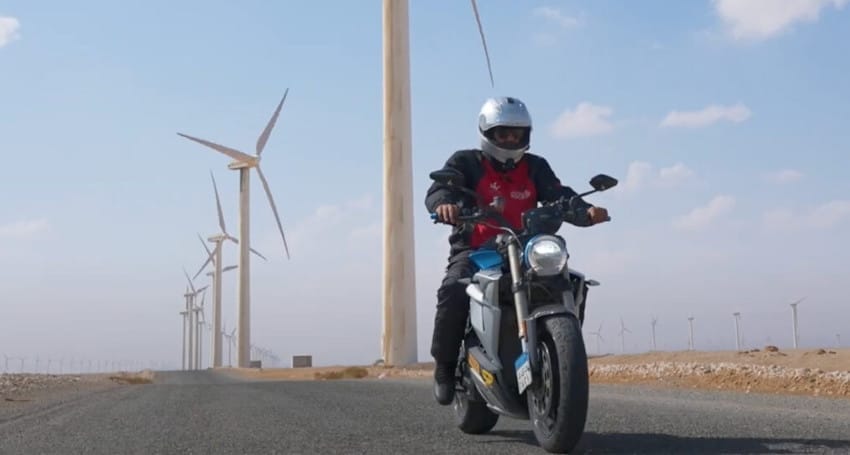 A person rides a motorcycle on a road beside wind turbines under a partly cloudy sky.