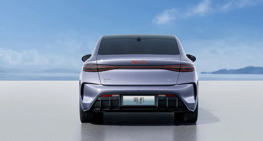 A silver car is shown from the rear, parked on a reflective surface, against a backdrop of a clear, blue sky and distant water and mountains.