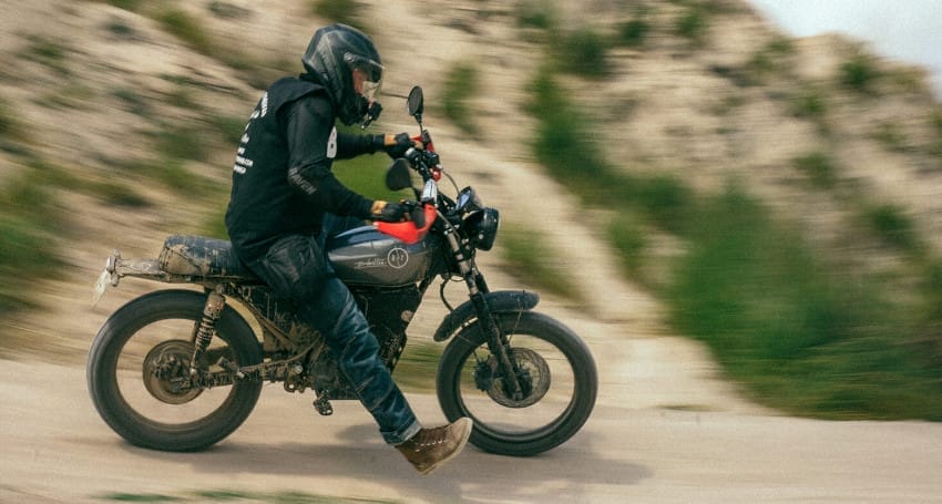 A person riding a Black Tea Motorcycles vintage motorcycle on a mountain road, shown in motion blur to emphasize speed.