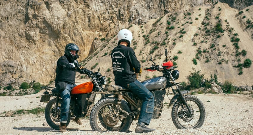 Two motorcyclists with helmets on dirt bikes discussing near a rocky hillside terrain, showcasing the vintage charm of their motorcycles.