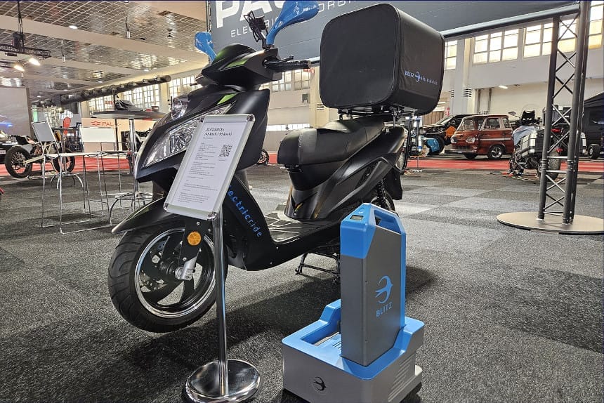 An electric scooter by Blitz Motors with a rear cargo box is displayed indoors on a carpeted floor, accompanied by an information stand and a removable battery placed beside the scooter. Ideal for eco-friendly last-mile deliveries, this setup suits services like Uber Eats perfectly.