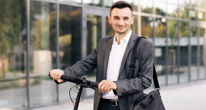 A smiling businessman in a suit and tie with a messenger bag, standing outdoors next to an electric scooter in front of a glass building.