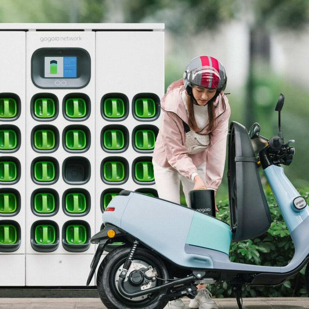 A woman is standing next to a green scooter in front of a charging station.