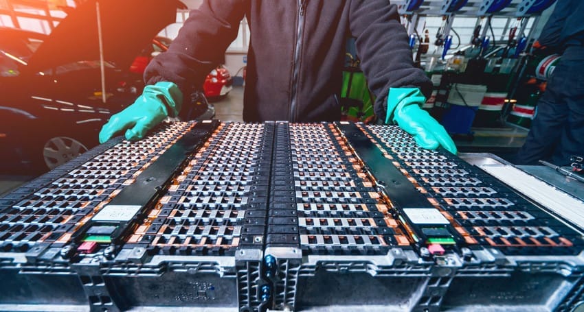 A person wearing green gloves is handling a large EV battery pack in an automotive workshop, with various tools and vehicles visible in the background.