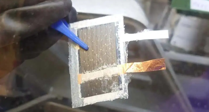 A person wearing a black glove holds a small rectangular solar cell with a blue tool. The solar cell has two attached metal strips, one silver and one copper.