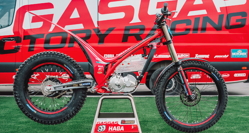 A red GASGAS TXE electric trial motorbike parked on a stand in front of the team van
