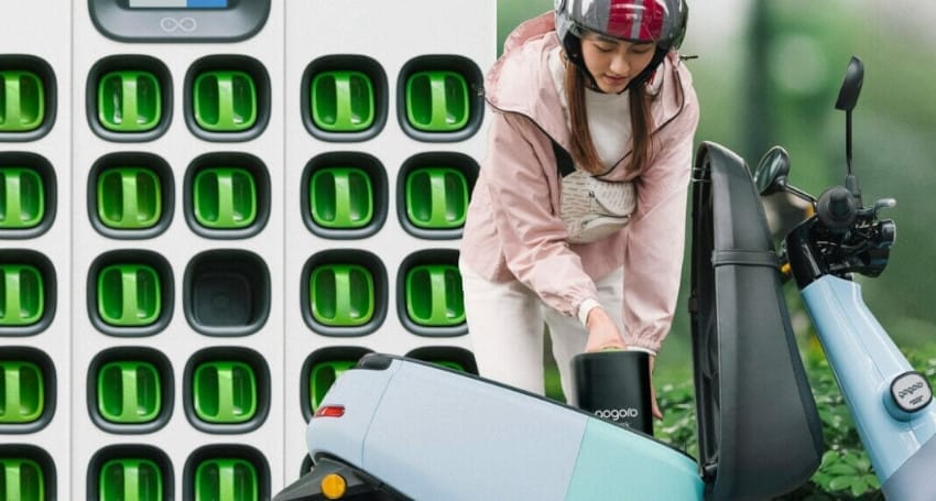 A woman riding a scooter next to an electric car charging station.