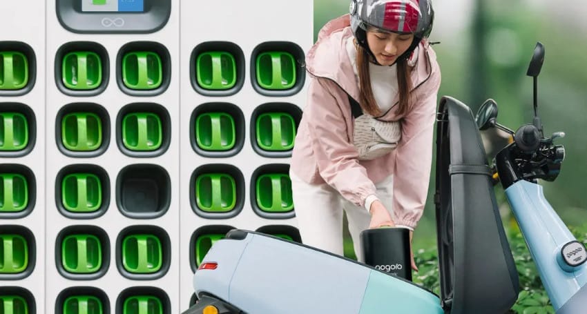 A person is swapping an EV battery in a light blue electric scooter next to a battery charging station filled with green batteries.