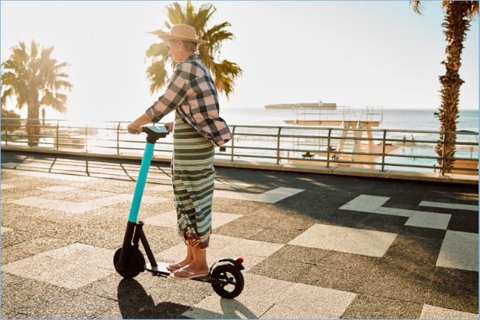 Woman in a straw hat and casual outfit riding an electric scooter along a sunny seaside promenade with palm trees.