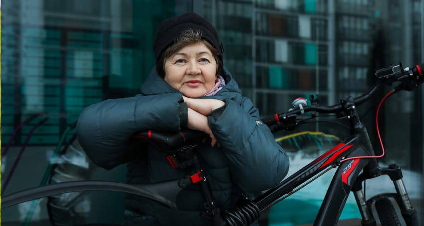 Mature woman in a winter coat and hat leaning on her best e-bike for your lifestyle, smiling, with a modern building in the background.
