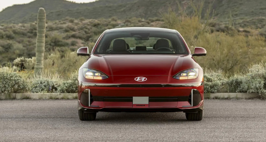 A red Hyundai IONIQ 6 electric car is parked on a paved road in front of a desert landscape with cacti and hills.