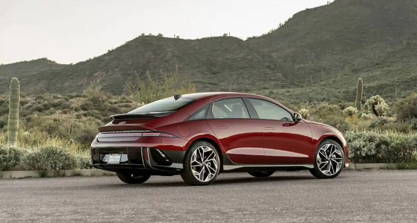 A red Hyundai IONIQ 6 sedan is parked on a road with desert vegetation and mountains in the background.