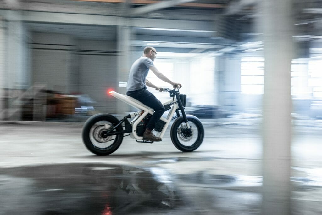 Adam Kay riding an electric Death To Petrol  motorcycle in a warehouse.