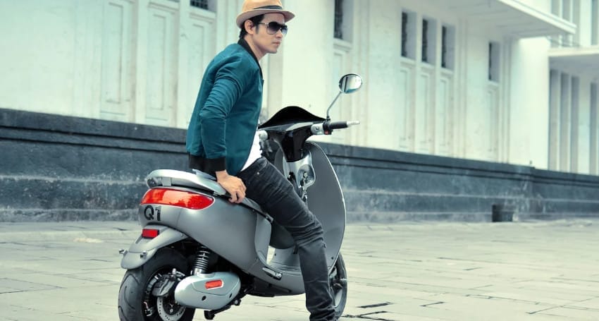A person wearing a hat and sunglasses sits on a grey scooter parked on a paved area outside a white building, showcasing the rise of electric motorbikes in Indonesia.