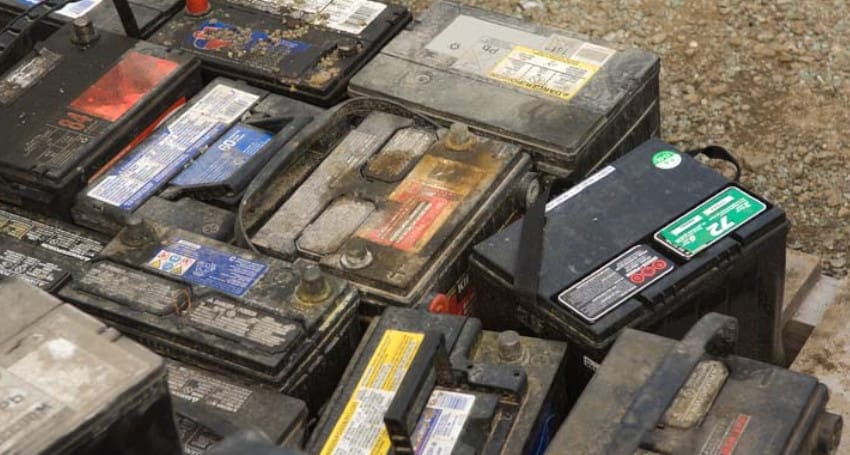 A collection of old and worn-out car batteries, including a few EV batteries, placed closely together, some with visible dirt and corrosion.