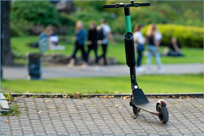 An electric scooter stands in the foreground on a cobblestone path with people walking in a blurred park background.