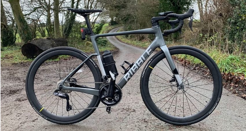 A grey Ribble bicycle, known as the lightest electric bike, with a black seat, black rims, and handlebars is parked on a narrow, tree-lined path.