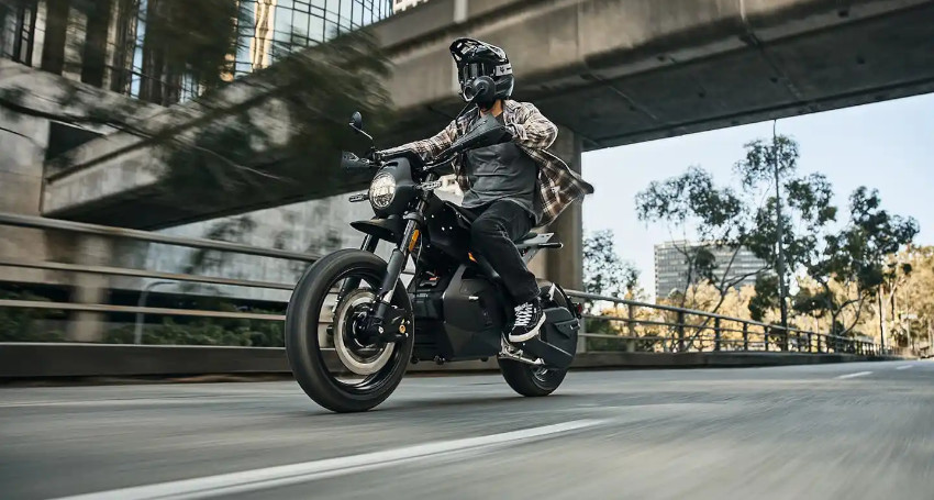 A person in a helmet rides a Ryvid Outset motorcycle on an urban road beneath an overpass on a clear day, with trees and buildings in the background.